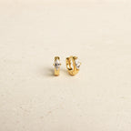 The Aurora Huggies are dainty small gold hoop earrings with marquise-shaped clear stones and subtle diamond accents, shown on a light beige surface.