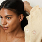 A woman with smooth skin wears Aurora Huggies, holding her dark hair back and dressed in a cream blouse with floral embroidery.