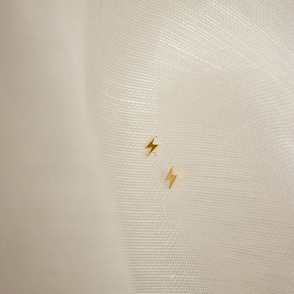 The Lightning Bolt Studs, featuring a small gold design, rest on a sheer white fabric background—perfect for adding an edgy jewelry accent to your look.