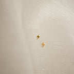 The Lightning Bolt Studs, featuring a small gold design, rest on a sheer white fabric background—perfect for adding an edgy jewelry accent to your look.