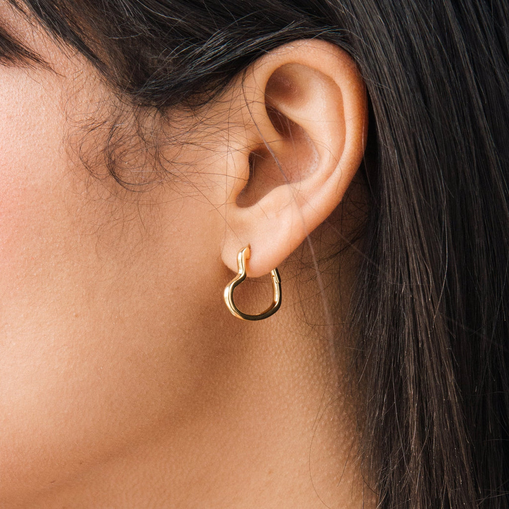 Close-up of a person wearing Heart Hoops, small gold heart-shaped earrings, with straight dark hair visible.