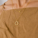 Woman in a brown dress wears two gold necklaces: the Pave Sun Necklace, featuring a sun pendant with pave accents, and another adorned with the letter N.