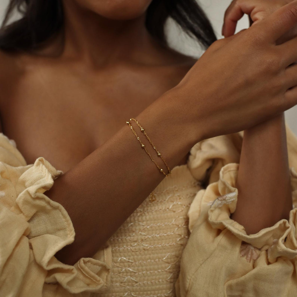 A woman in a cream ruffled dress wears a dainty gold chain and the Duo Bead Chain Bracelet in Sterling Silver on her wrist.