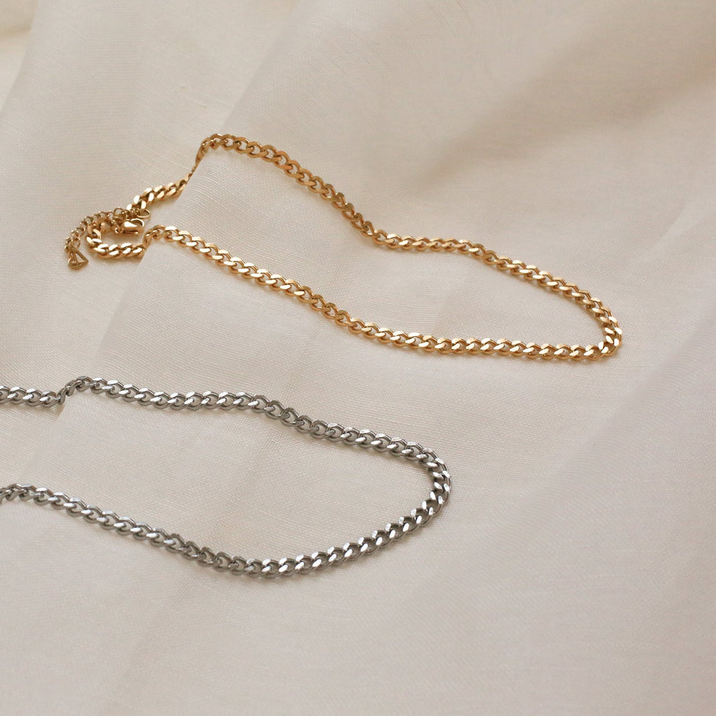 A flat lay showing both the gold and silver versions of the curb chain necklace on a cream fabric.