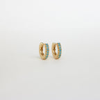 The Kendra Huggies are small gold hoop earrings accented with a row of turquoise stones, adding eclectic charm to any style and shown on a plain white background.