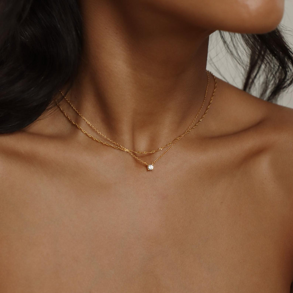 A woman wears the Dainty Diamond Necklace, a double-layered gold piece featuring a subtle diamond charm resting at the base of her neck, with her bare shoulders and collarbones showing.