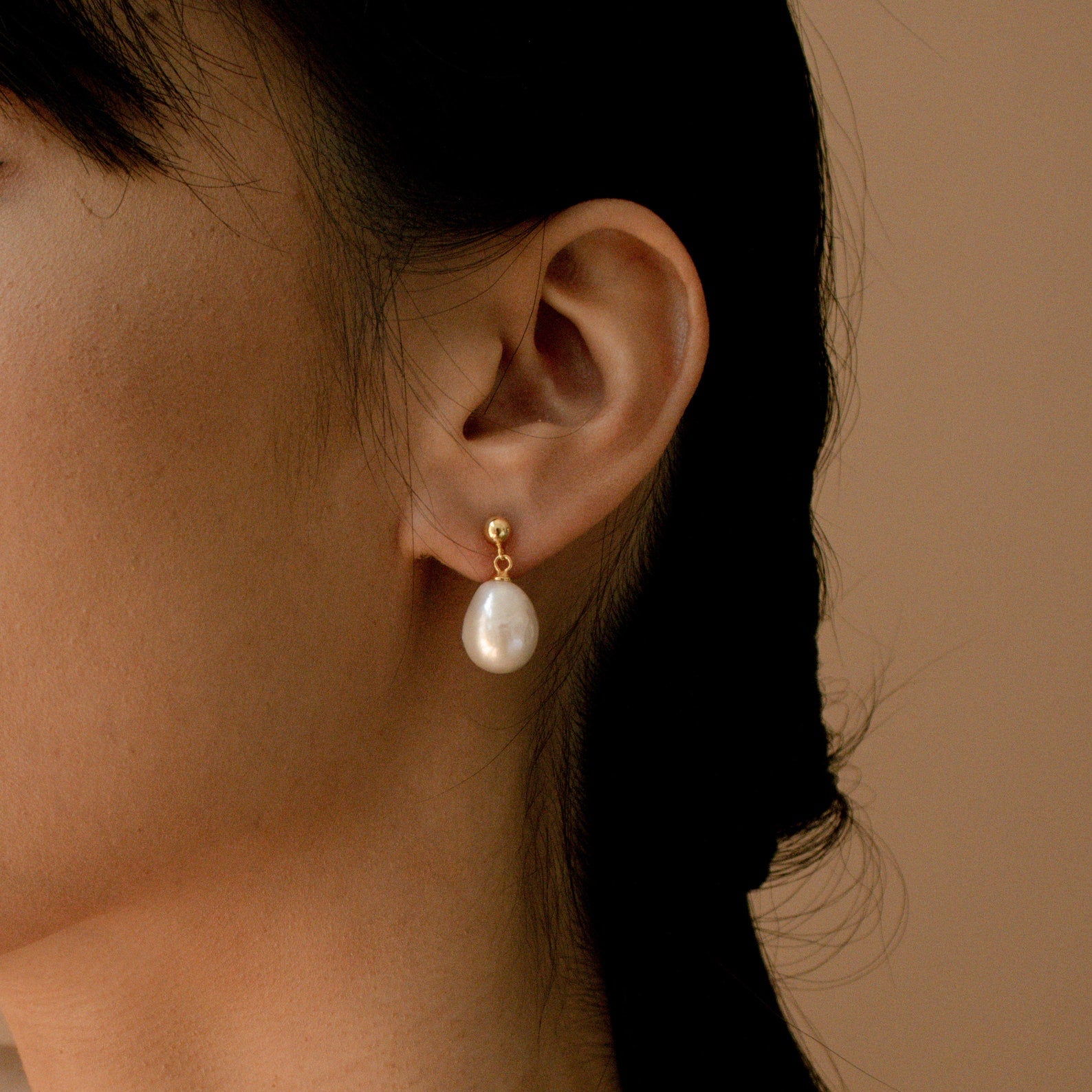 Close-up of a woman wearing Baroque Pearl Earrings with gold studs and large baroque pearl drops, set against a beige background.