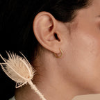 Close-up of a woman's ear wearing Parker Pave Hexagon Huggies, gold geometric earrings, with a dried thistle flower in the foreground.