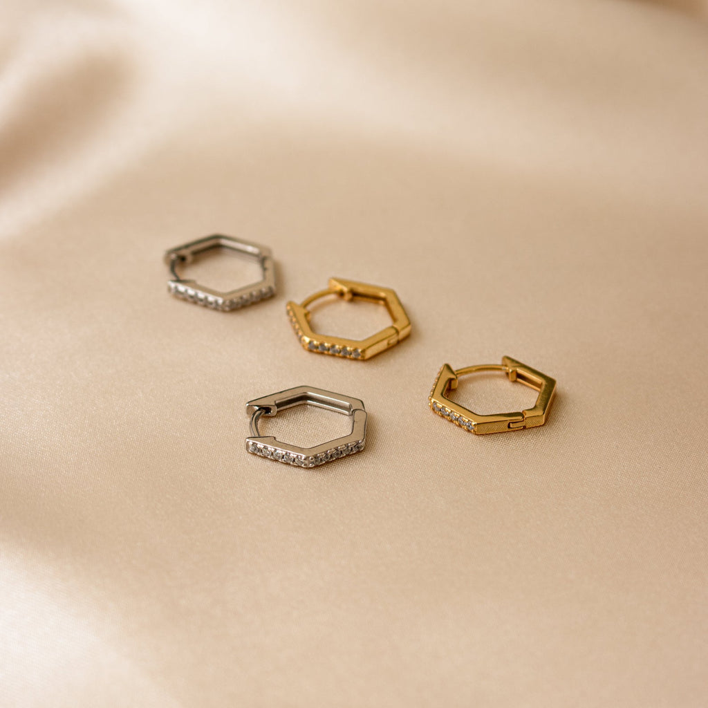 Two pairs of Parker Pave Hexagon Huggies—one silver, one gold—featuring small crystals for sparkle, displayed on beige fabric. A chic ear accessory.