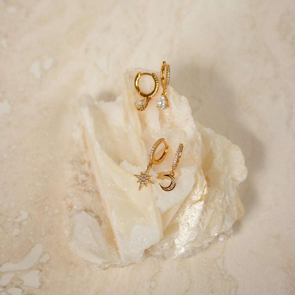 The Moon and Star Huggies in Sterling Silver, featuring celestial-inspired designs with crystal accents, are displayed on a white translucent rock against a beige background.