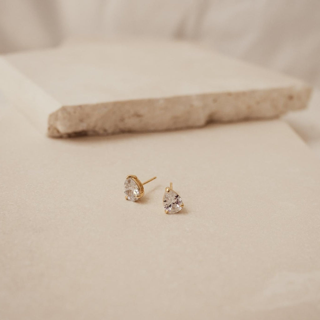 The Pear Diamond Studs, crafted in gold with a teardrop shape, rest on beige stone tiles, highlighting the elegant appeal of minimalistic earrings.