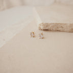 The Oval Diamond Studs are classic gold earrings featuring clear gemstones, displayed on a beige stone surface.