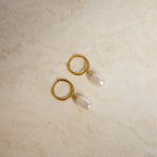 The Maggie Pearl Drop Hoops feature modern oval pearls hanging from sleek hoops, shown on a light beige, textured surface—ideal for minimal jewelry lovers.