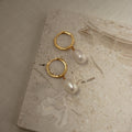 Maggie Pearl Drop Hoops—gold hoops with dangling white pearls—are shown on a light textured stone surface, making them the perfect minimal jewelry piece.