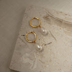 Maggie Pearl Drop Hoops—gold hoops with dangling white pearls—are shown on a light textured stone surface, making them the perfect minimal jewelry piece.