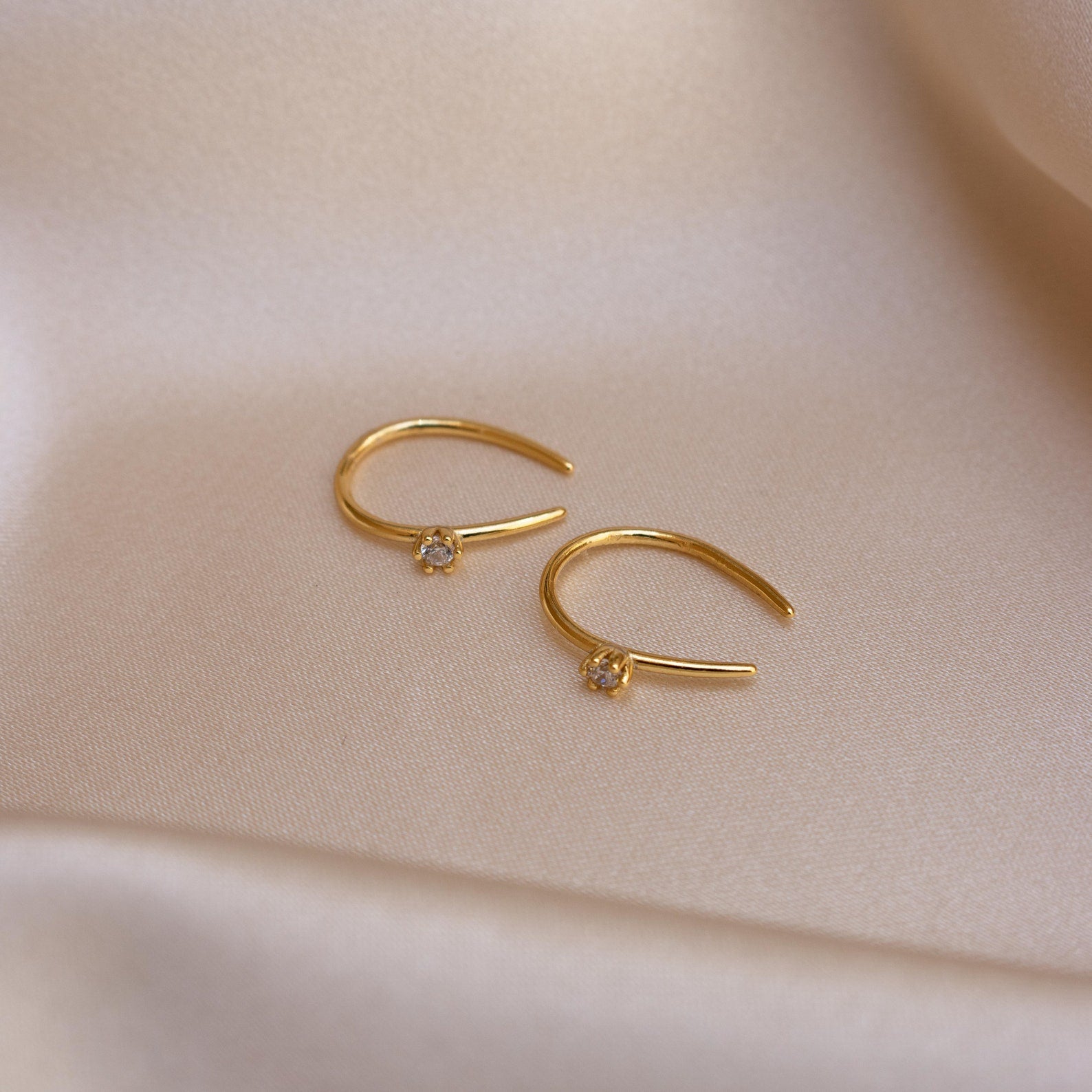 The Rylee Diamond Wire Earrings, featuring two gold open hoops set with small diamonds, rest on soft beige satin fabric, adding a modern touch of sparkle.