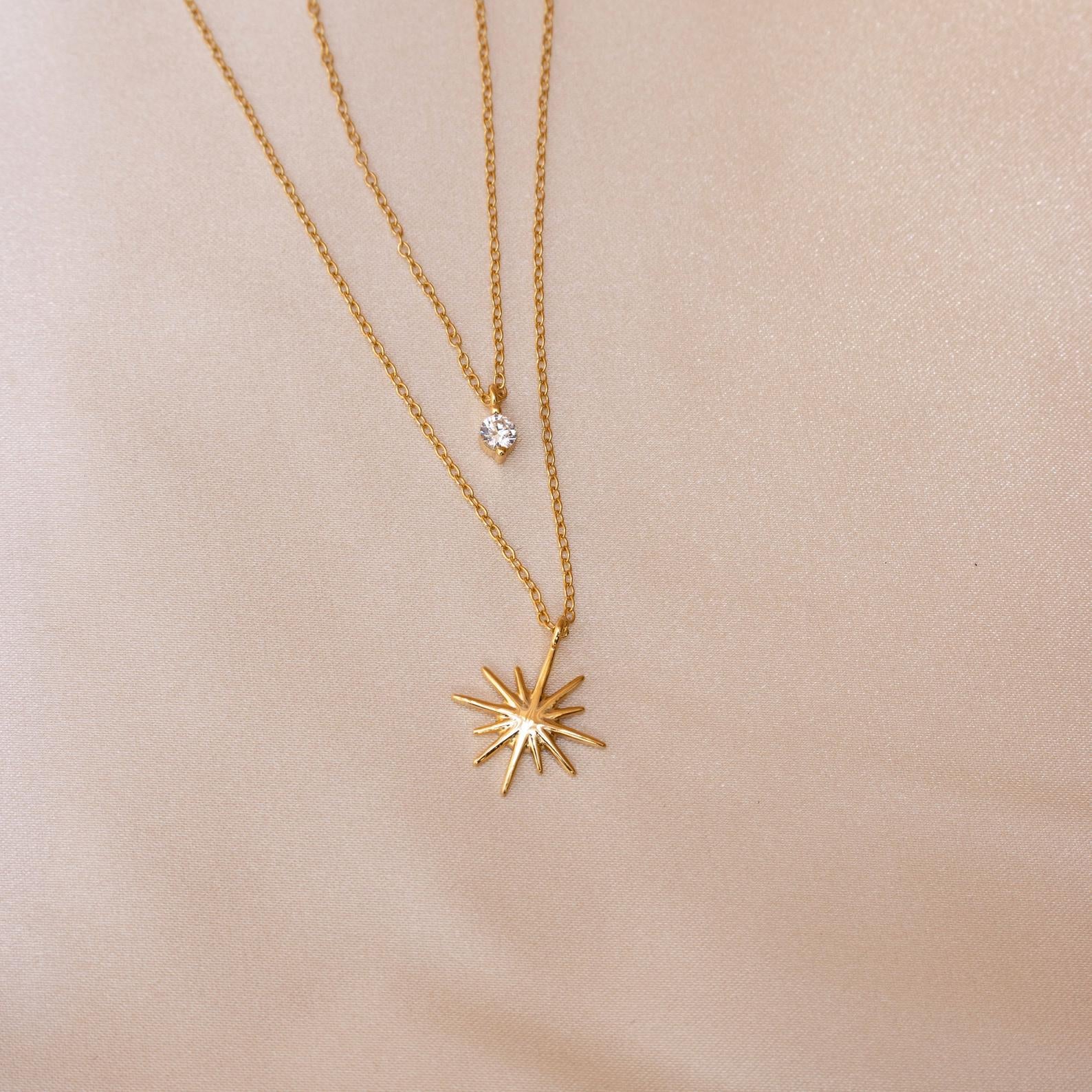 The Laura Starburst Duo Necklace in 18K Gold is displayed on light beige fabric, featuring two gold chains—one with a diamond-shaped pendant and the other with a starburst pendant set with a central gemstone.