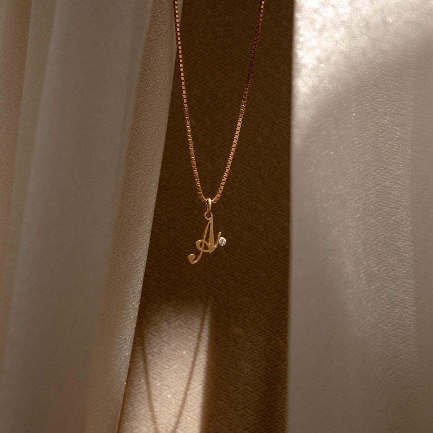 The Initial Birthstone Necklace features an A pendant and a sparkling gemstone, beautifully displayed between soft beige curtains—a sophisticated personalized gift and elegant custom monogram charm.