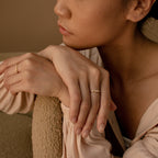 A woman rests her chin on her hands, highlighting a slim Baguette Diamond Ring and gold rings, styled with a beige blouse against a neutral backdrop.
