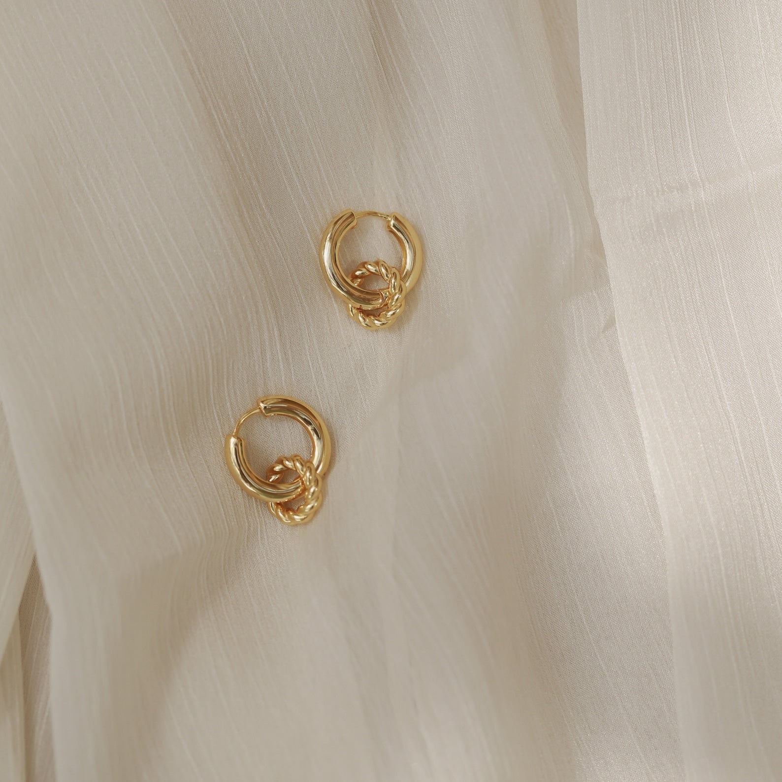 The Claire Duo Hoops are chunky earrings with elegant knot details, shown on sheer light beige fabric—ideal for fans of chic jewelry.