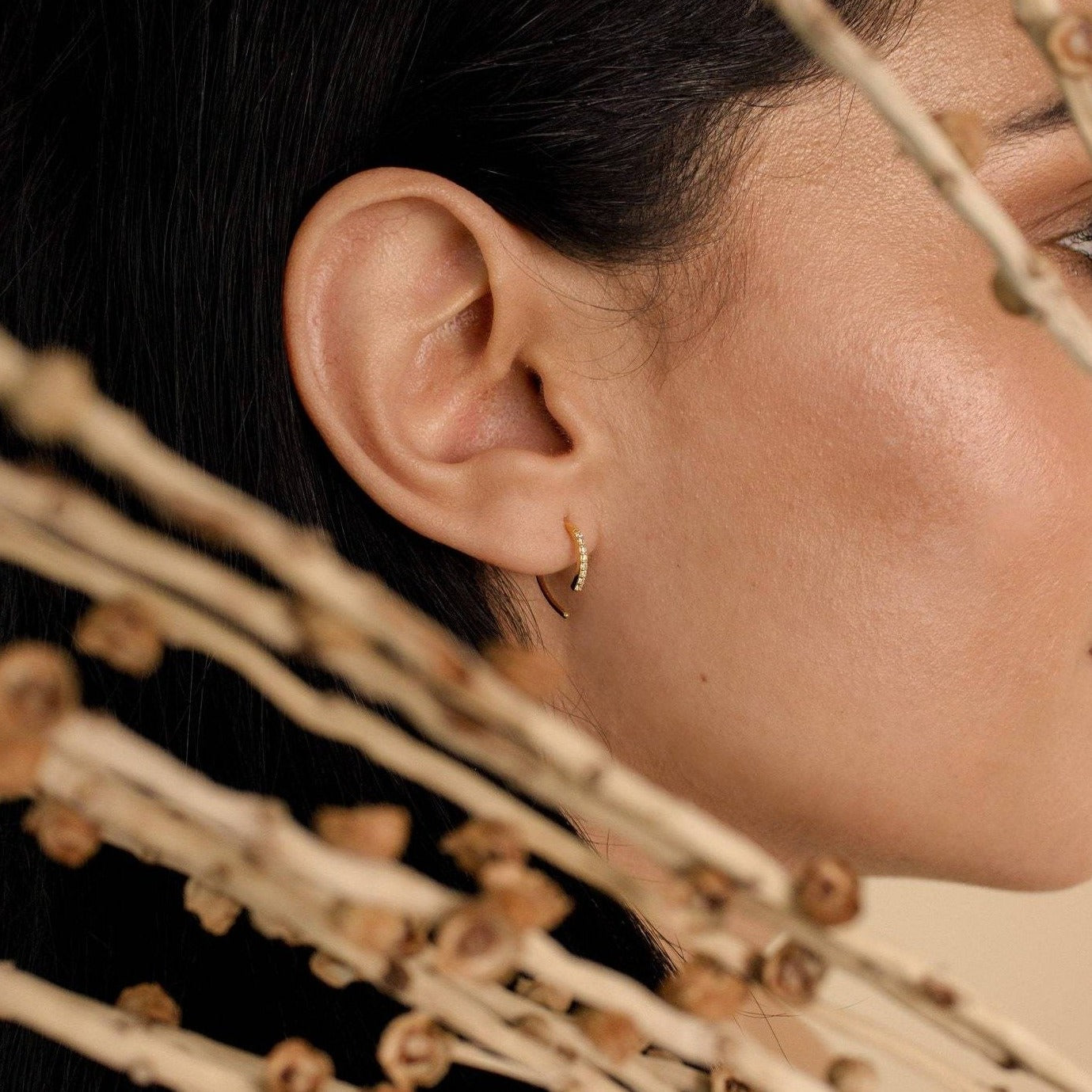 Close-up of a woman's ear wearing Pave Wire Diamond Hoops in gold, with dried twigs in the foreground.