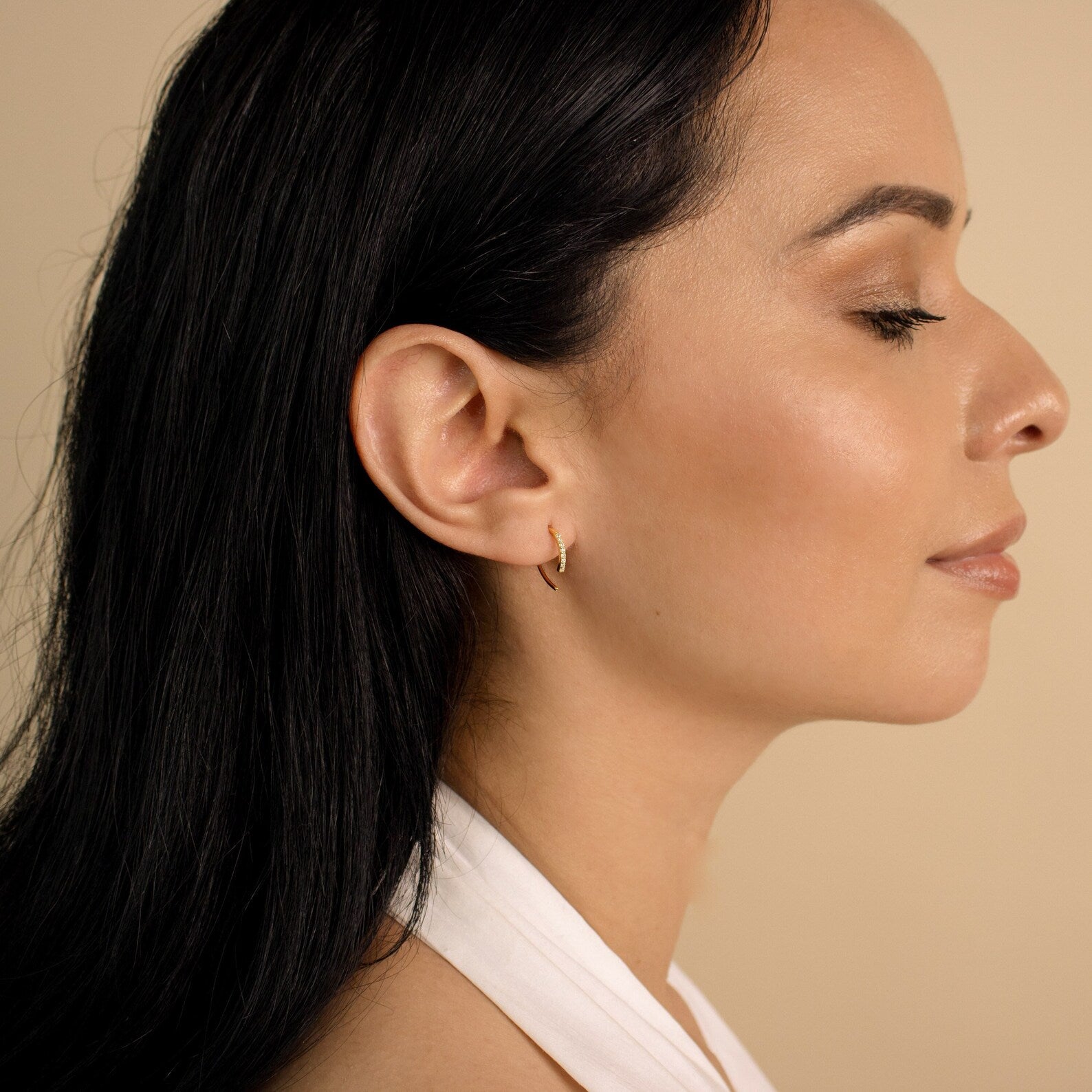A woman with long dark hair, wearing Pave Wire Diamond Hoops, faces right with eyes closed against a beige background.