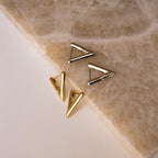 Two pairs of Triangle Huggies, in gold and silver finishes, are displayed on a textured stone surface—an ideal accessory for effortless summer style.