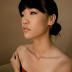 A woman with dark hair in a low bun, wearing Medium Thin Hoops, gazes thoughtfully to the side.