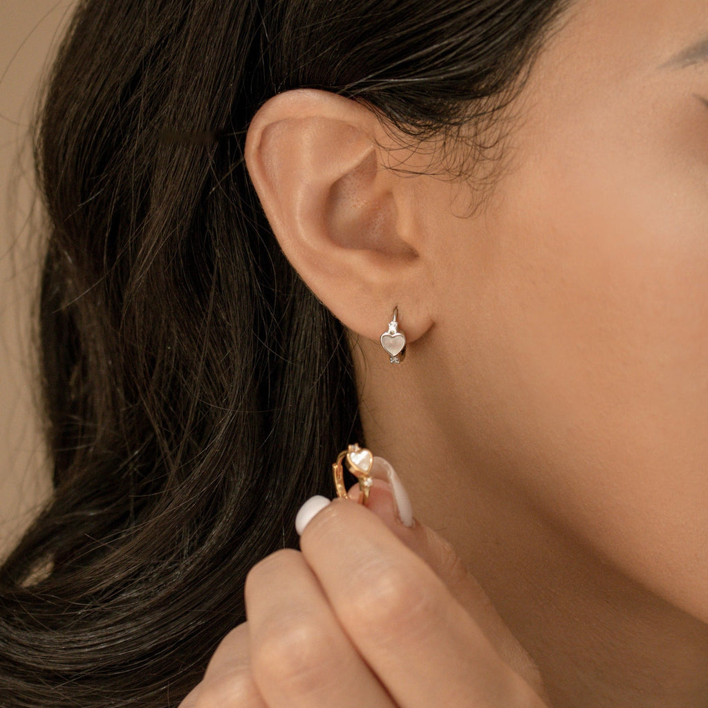 Close-up of a woman wearing a small heart-shaped earring and holding another Scarlett Pearl Huggies earring near her ear.