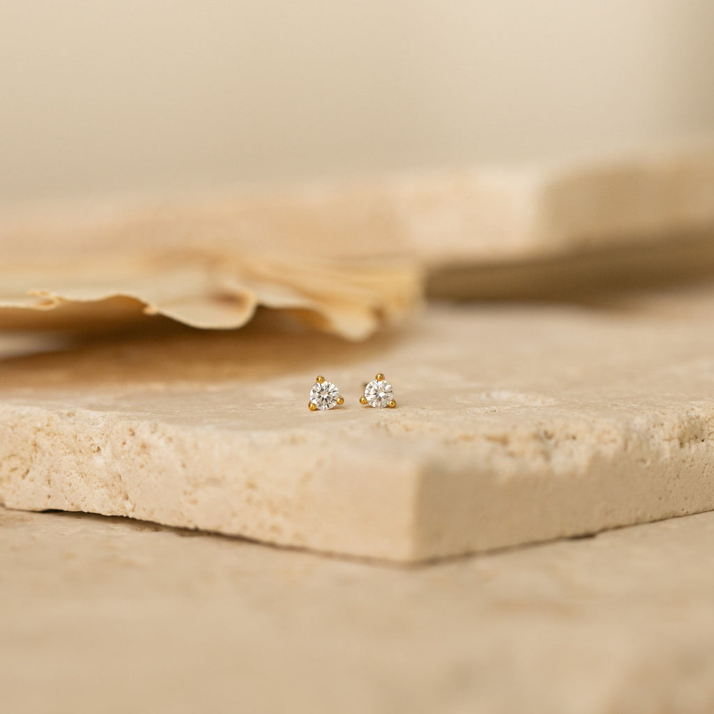 The Elena Diamond Studs feature three clustered diamonds set in small gold studs, resting elegantly on a textured beige stone—perfect minimalist earrings for everyday wear.