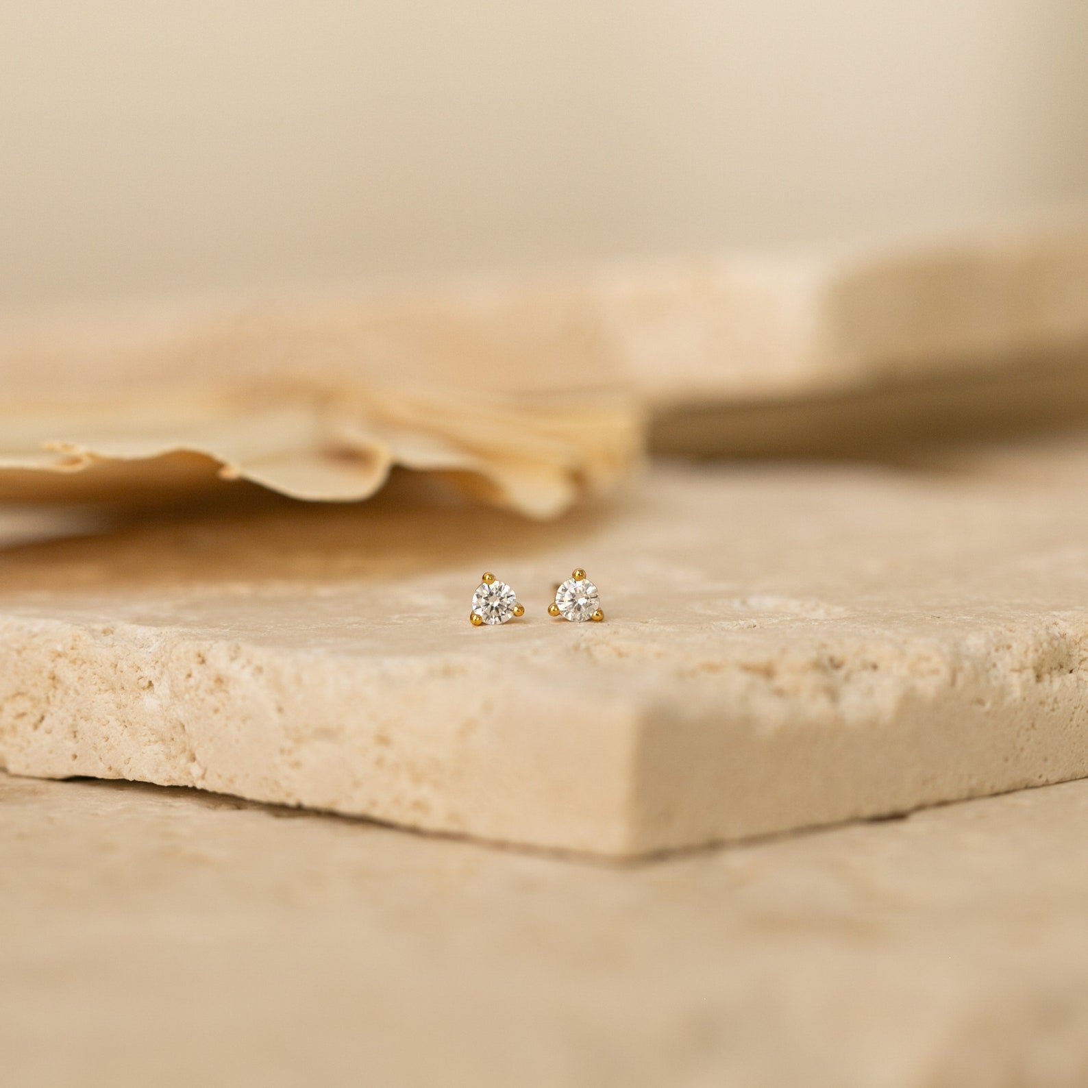The Elena Diamond Studs feature three clustered diamonds set in small gold studs, resting elegantly on a textured beige stone—perfect minimalist earrings for everyday wear.