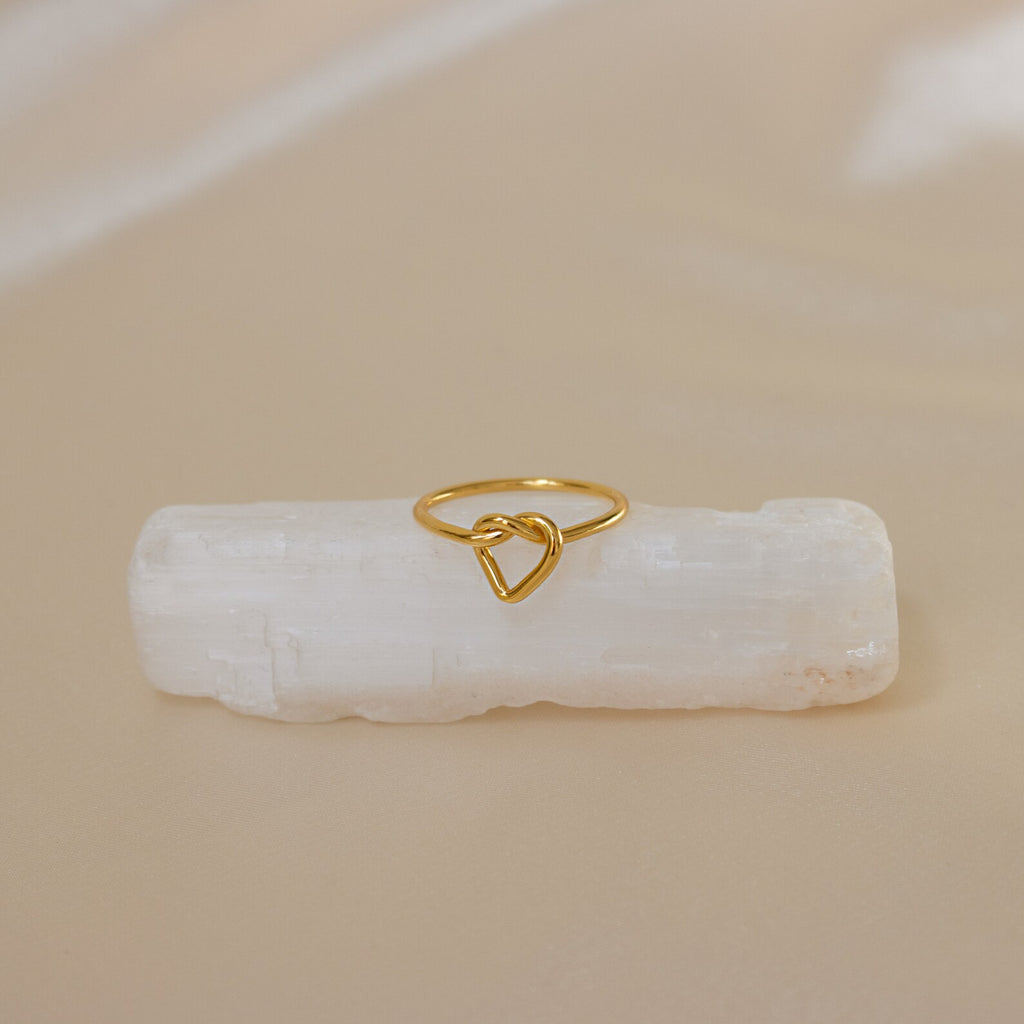 The Love Heart Knot Ring features a heart-shaped knot design, displayed on a white crystal with a beige background—a delicate and dainty piece ideal as a promise ring.