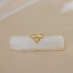 The Love Heart Knot Ring features a heart-shaped knot design, displayed on a white crystal with a beige background—a delicate and dainty piece ideal as a promise ring.