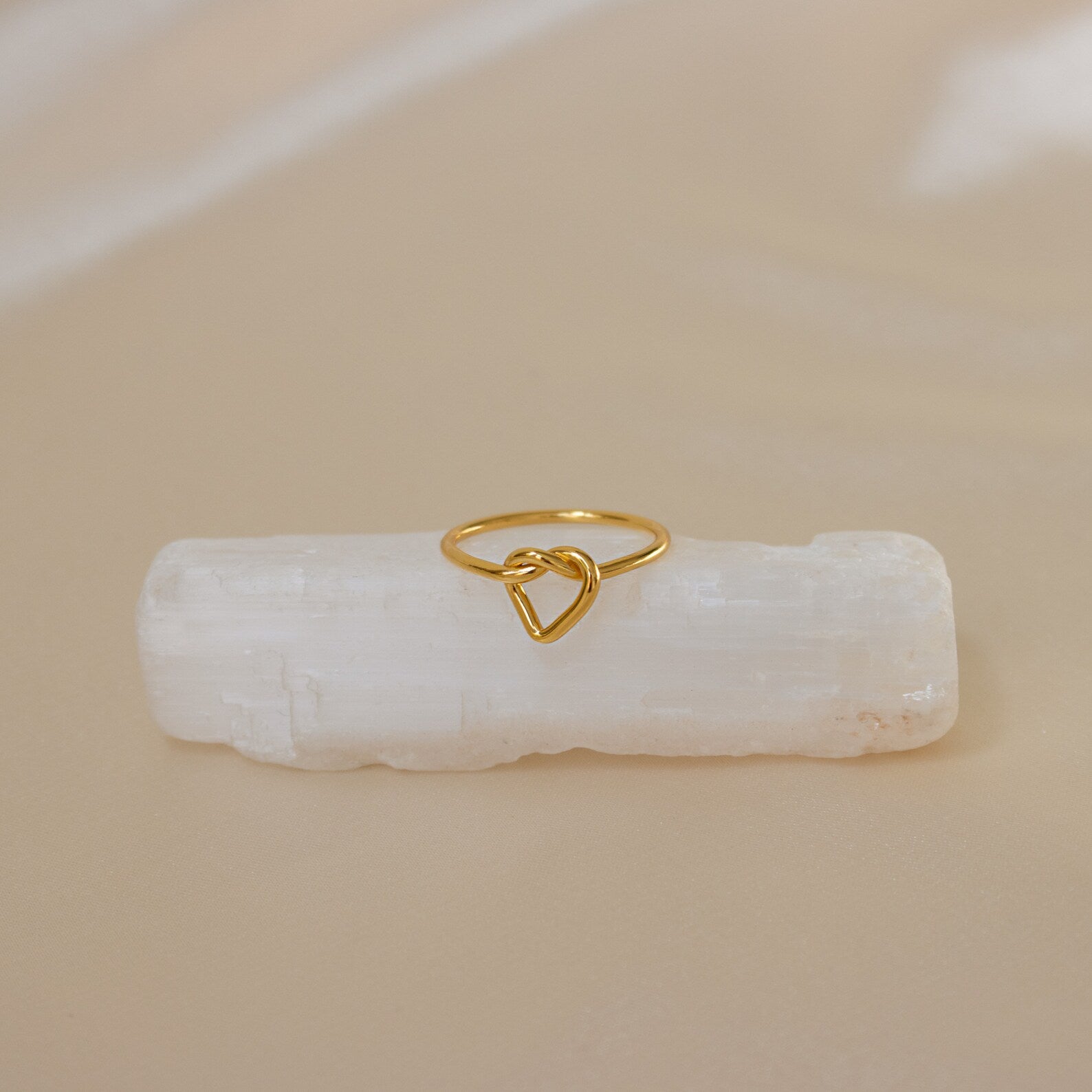 The Love Heart Knot Ring features a heart-shaped knot design, displayed on a white crystal with a beige background—a delicate and dainty piece ideal as a promise ring.