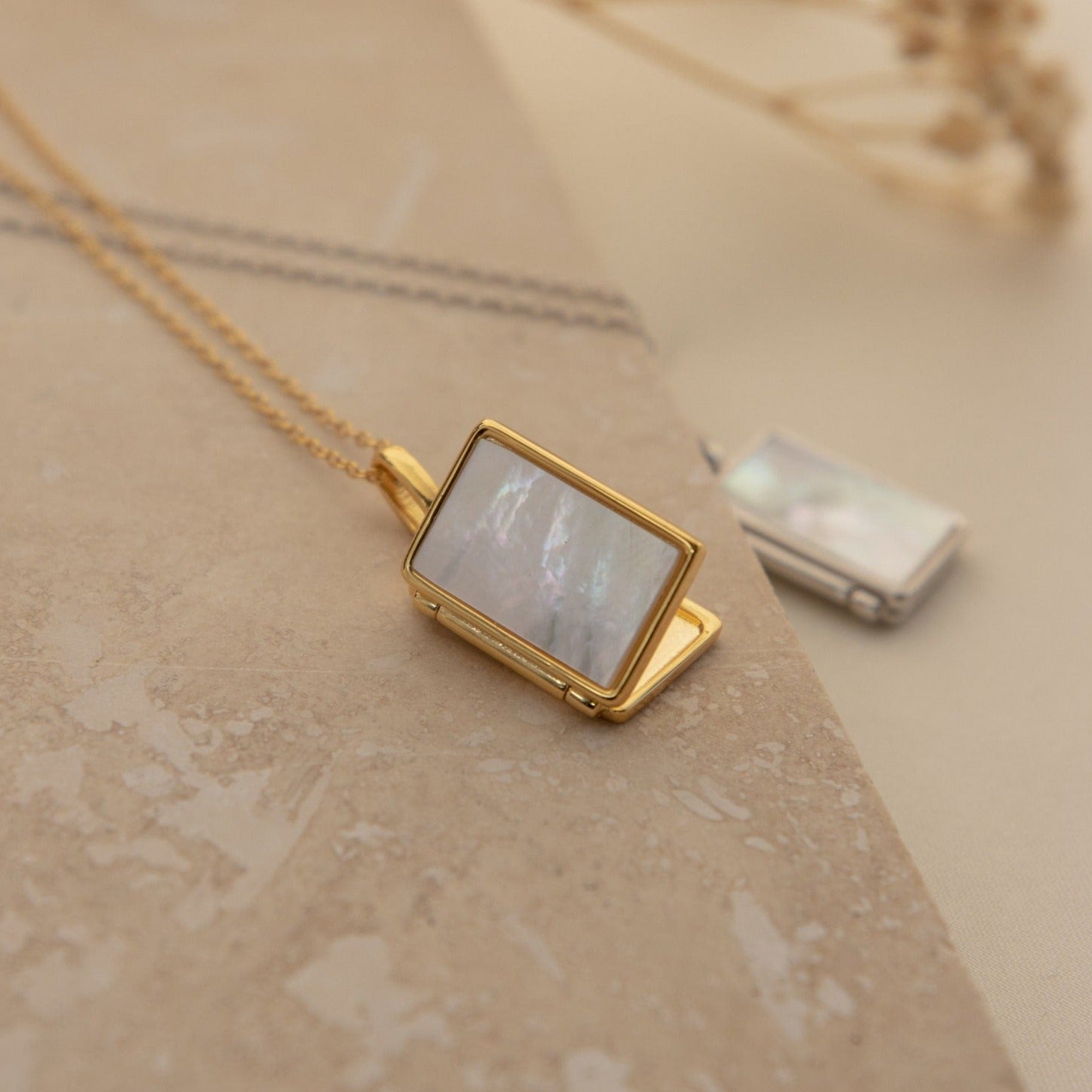 A gold rectangular vintage Pearl Locket Necklace with a Mother of Pearl inlay rests on a beige stone surface, while another silver Pearl Locket Necklace is softly blurred in the background.