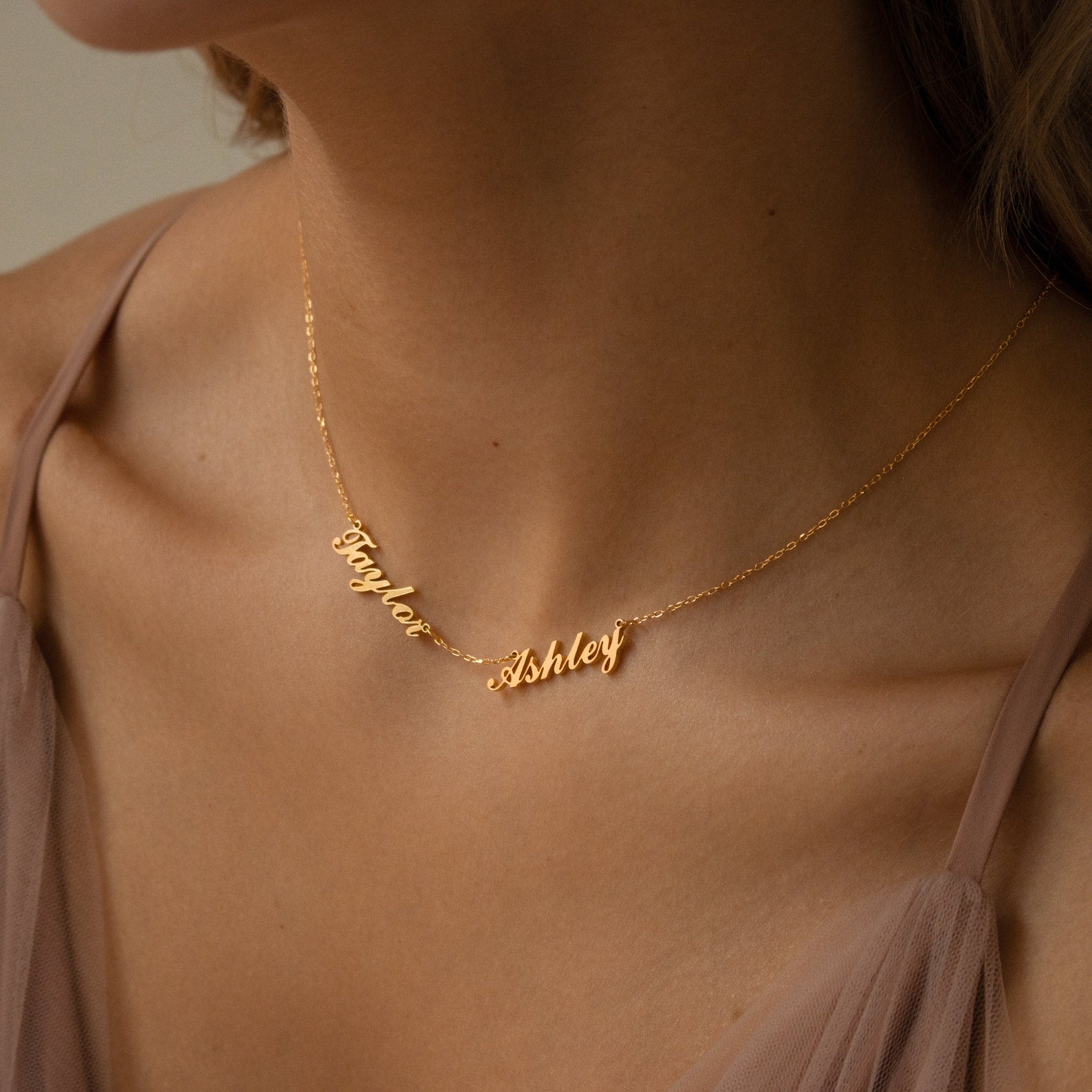 The Carrie Multiple Name Necklace in Rose Gold, featuring “Taylor Ashley” in elegant script, rests gracefully on a woman's neck and complements her sheer blush top—a beautiful example of personalized jewelry.