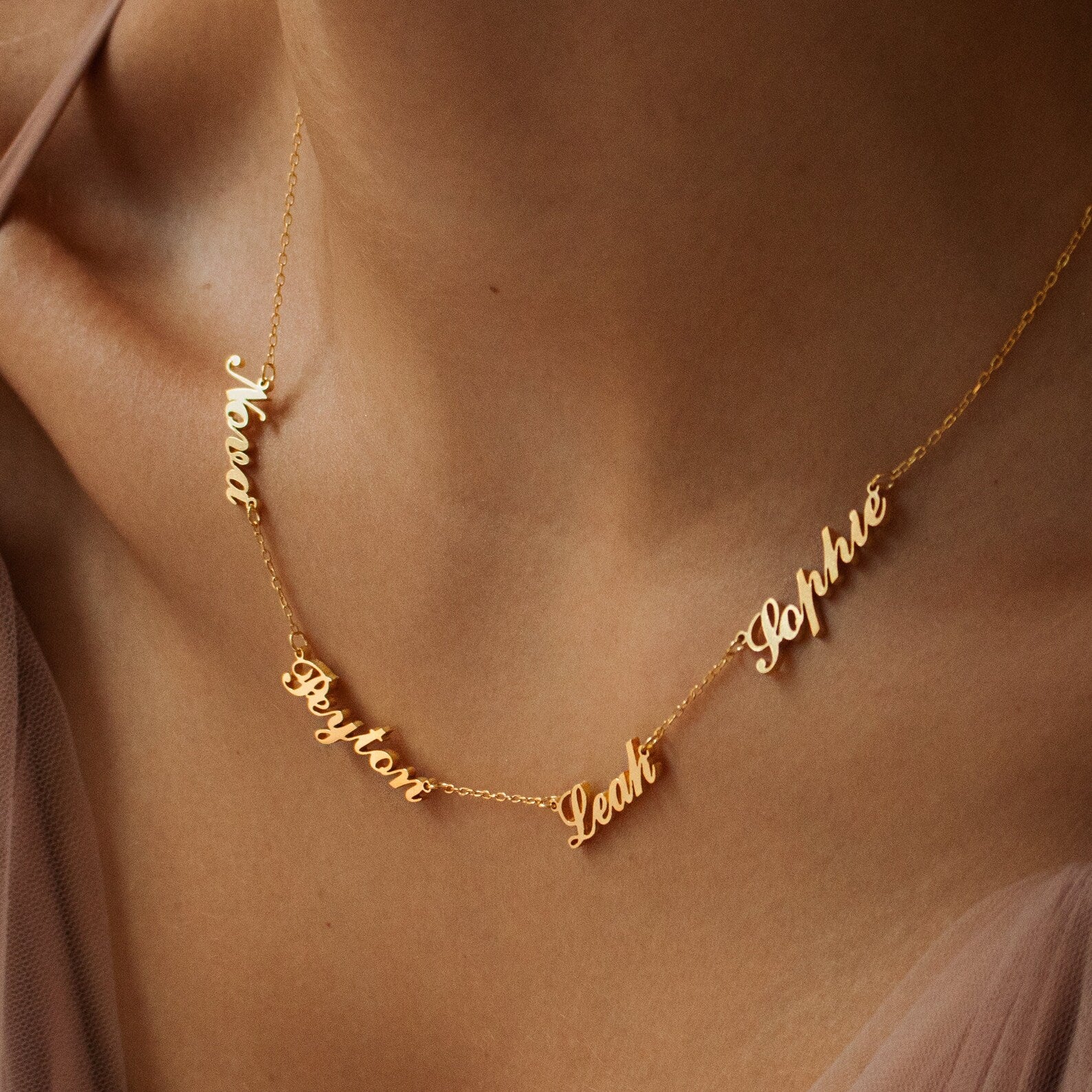The Carrie Multiple Name Necklace in Rose Gold features a delicate chain and multiple names—Noah, Peyton, Leah, and Sophie—elegantly scripted in cursive on a woman’s neck.