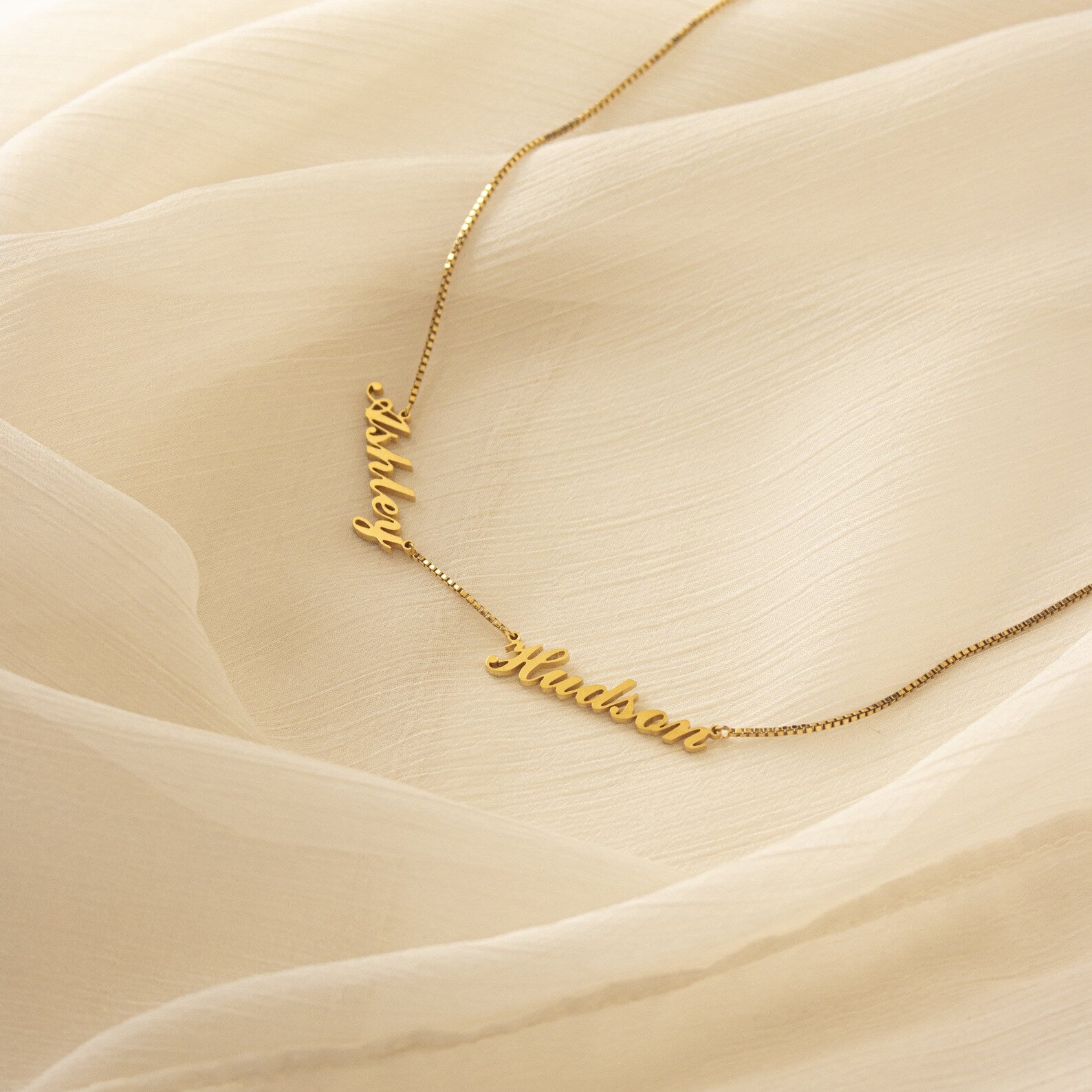 Carrie Multiple Name Necklace in Box Chain, shown with "Ashley" and "Hudson" on a cream sheer fabric background—perfect personalized jewelry for a unique touch.