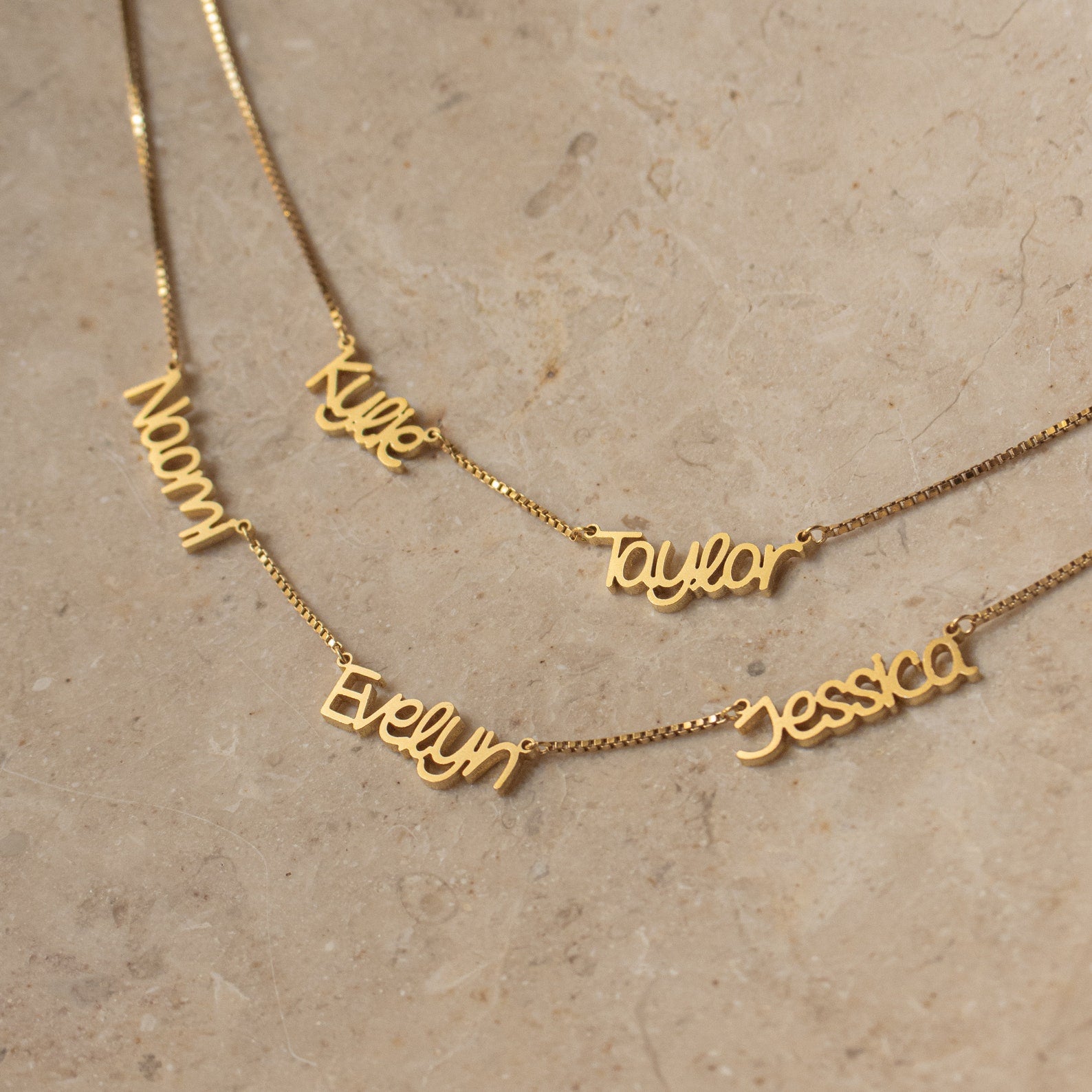 Showcasing the Preppy Multiple Name Necklace in Box Chain: five gold necklaces with personalized names—Naomi, Evelyn, Kylie, Taylor, and Jessica—in a chic youth-inspired font on a beige stone surface.
