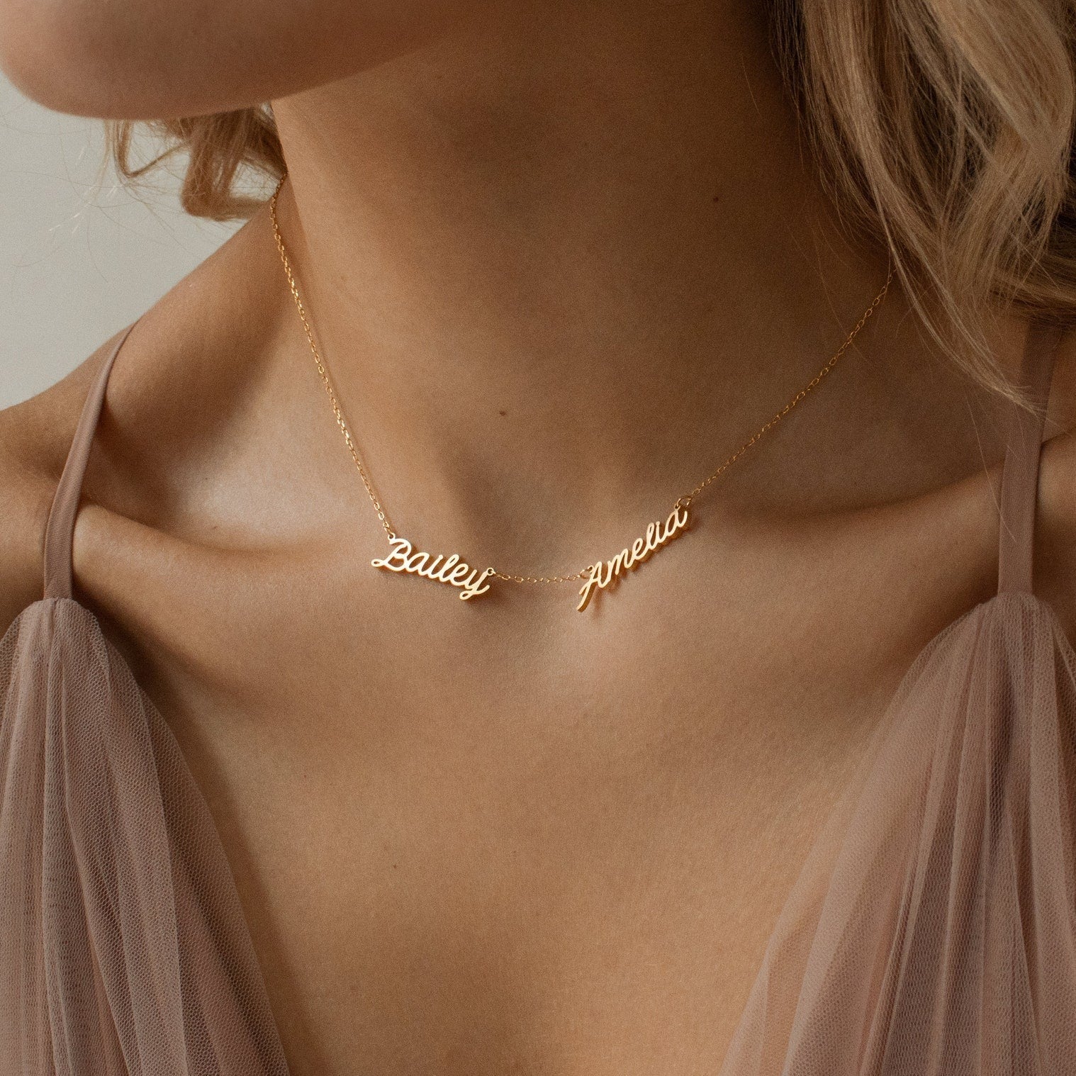 Close-up of a woman wearing the Paris Multiple Name Necklace in gold with Bailey and Amelia pendants, styled with taupe fabric.