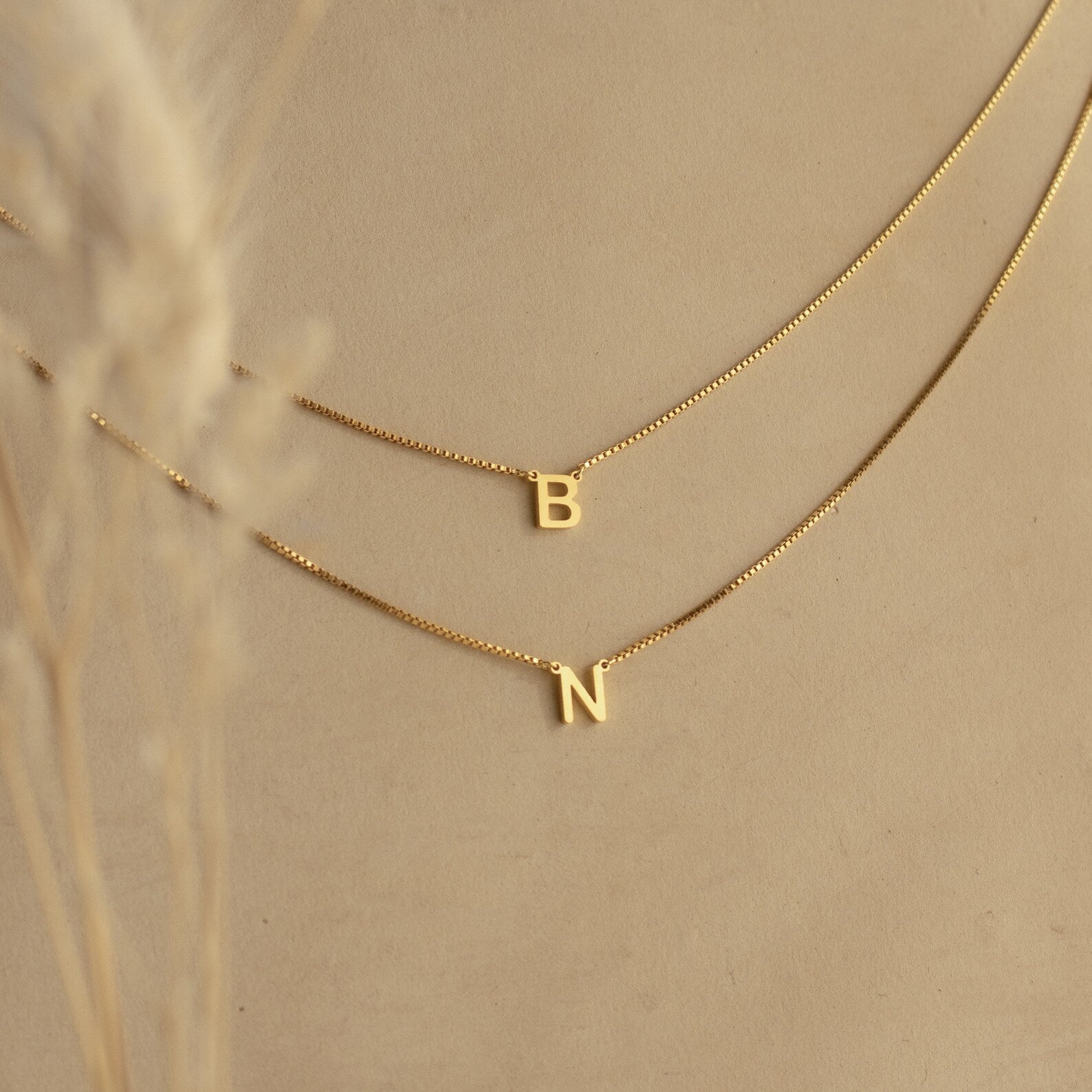 The Alice Double Letter Necklace in Box Chain showcases gold letter pendants B and N on a beige background—a stunning set for personalizing your style.