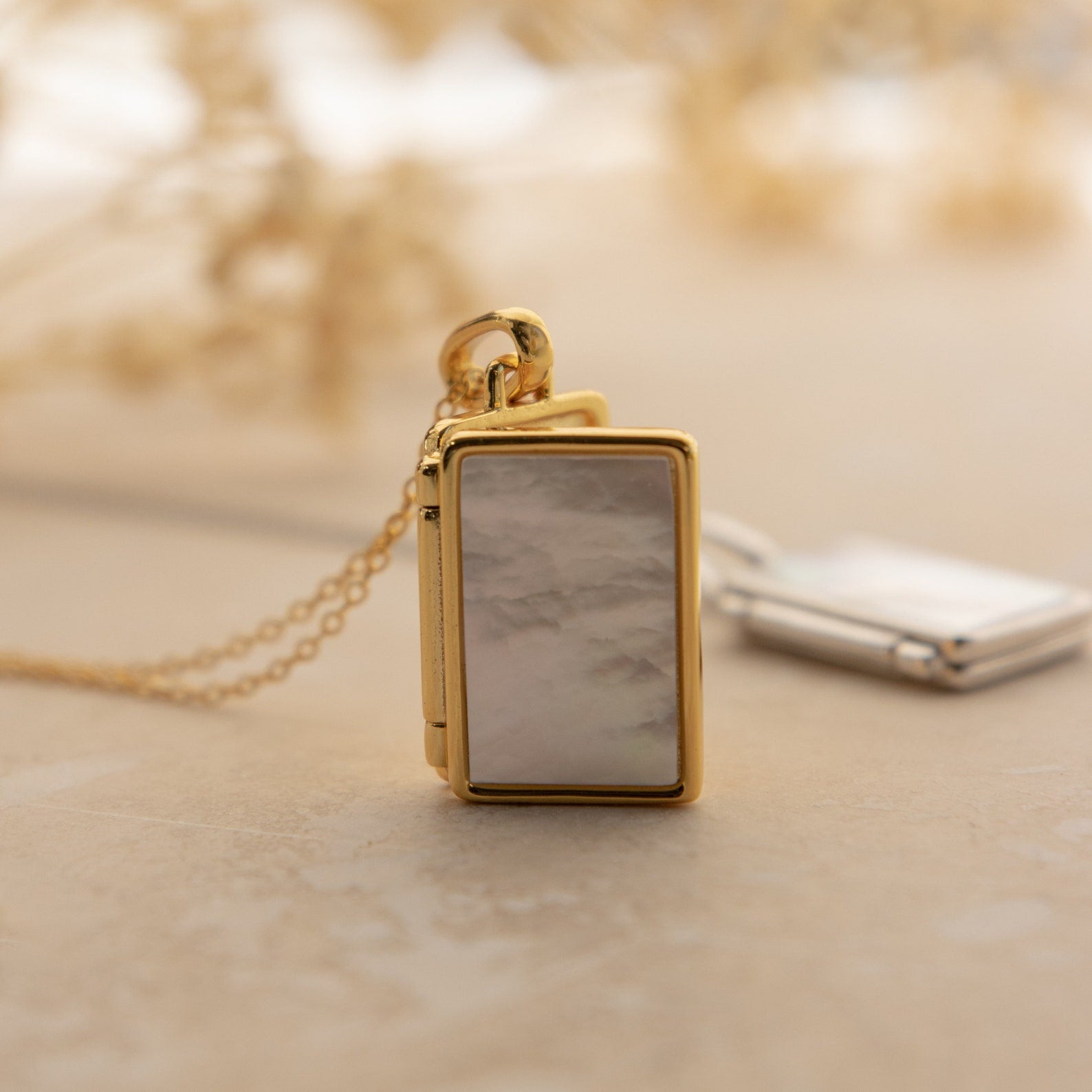 Close-up of a rectangular gold locket necklace with a glossy mother-of-pearl inlay, resting on a stone surface with a blurred silver version in the background.