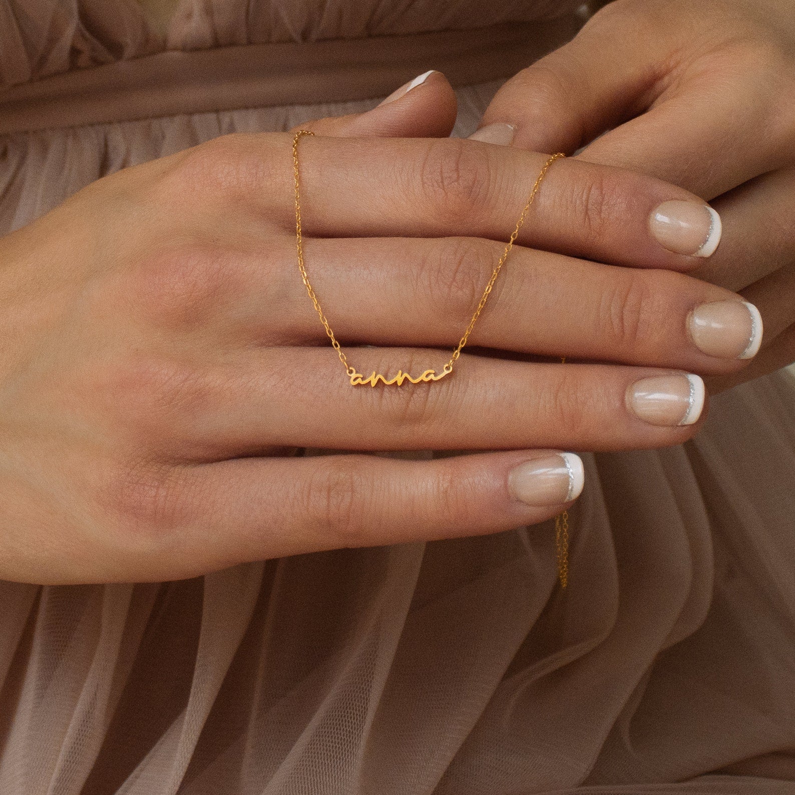 Close-up of hands holding the Tiny Summer Name Necklace in a delicate font with the name "anna." The wearer is dressed in a sheer beige dress, highlighting the necklace’s elegant and personalized design.