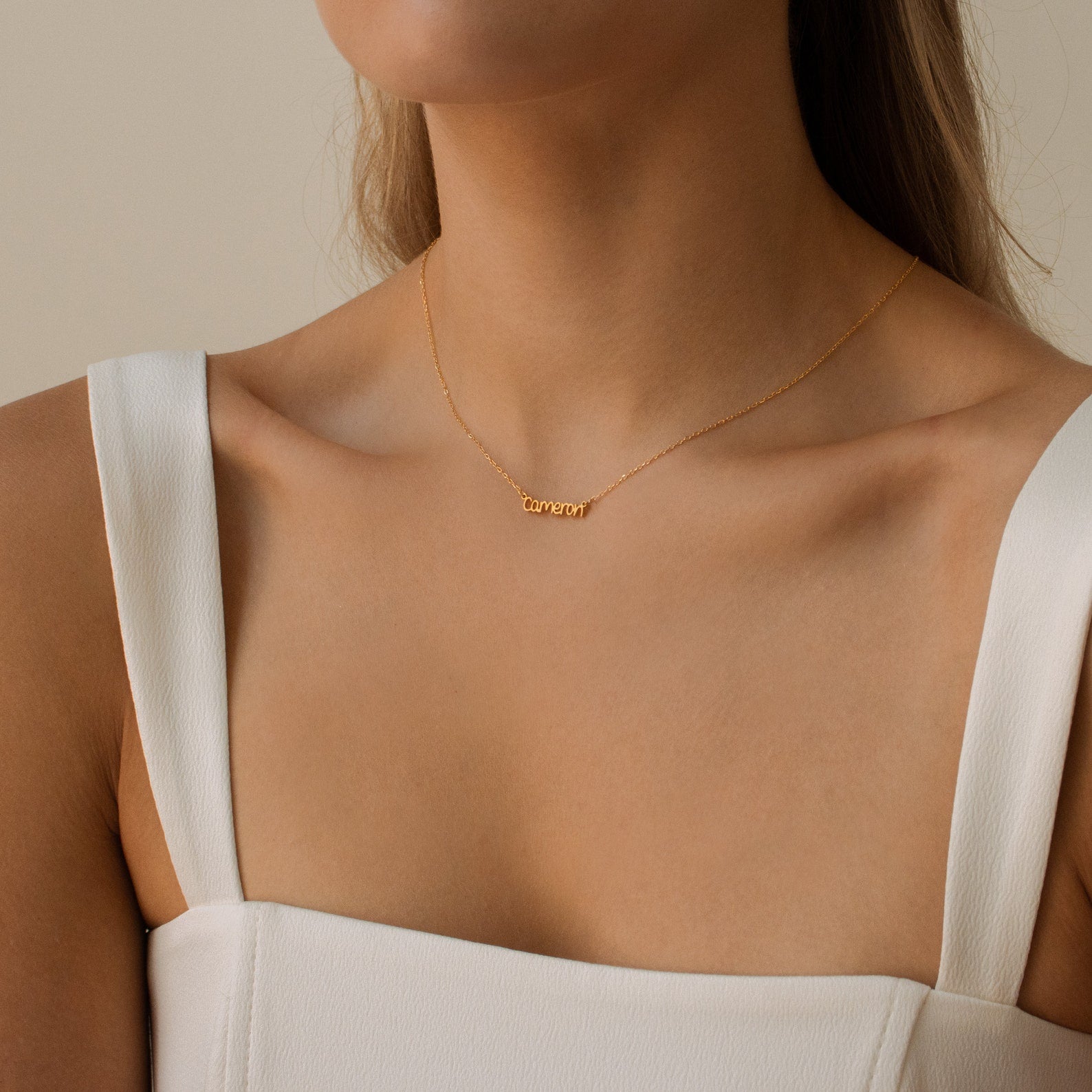 A woman models the Tiny Preppy Name Necklace in gold, customized with the word "consent" in a youth-inspired font, paired with a white sleeveless top and shown from the shoulders up.