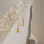 Two Nella Letter Necklaces rest on a textured white stone, framed by dried foliage—perfect for layering or gifting as a personalized accessory.