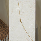 The Pave Sideways Initial Necklace, featuring a gold chain and small vertical rectangular charms, is displayed on a light stone background.