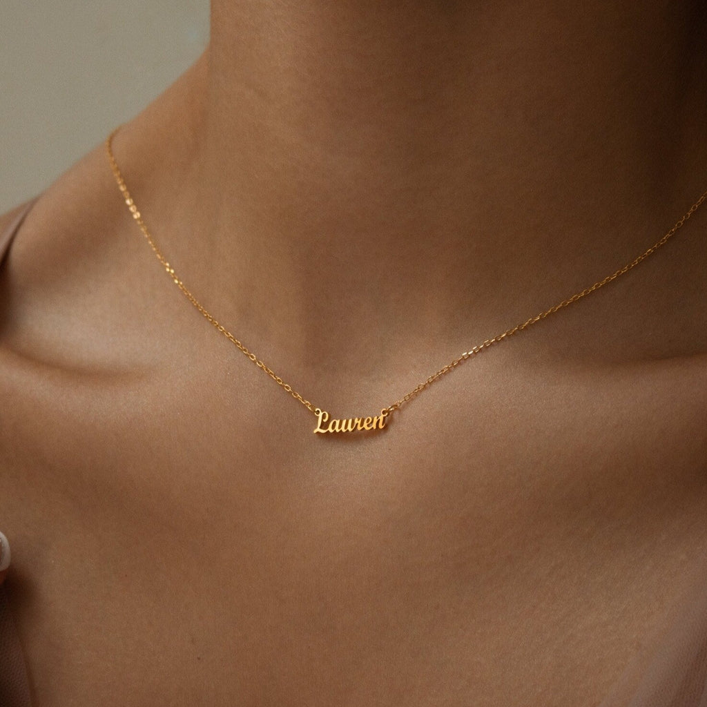 The Tiny London Name Necklace features a customizable name plate resting on the neck and collarbone, offering a chic, minimalist style.