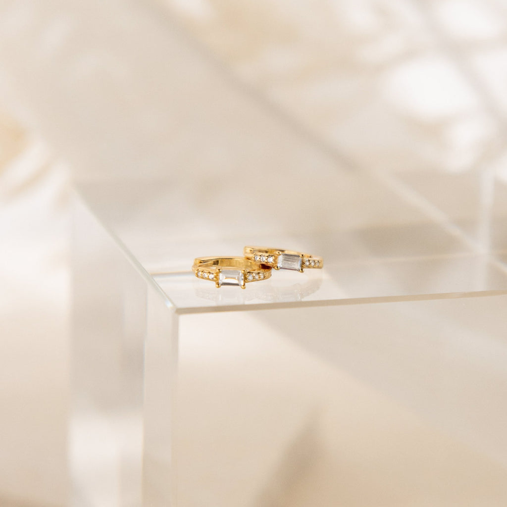 The Natalie Pave Huggies, with sparkling gemstones, are displayed on a glass-like surface against a soft background, echoing the elegance of baguette diamond and dainty hoop earrings.