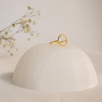 The Pave Open Circle Ring features a gold band set with small gems, showcased on a smooth white dome with flower sprigs in the background for an elegant touch.