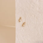 The Nile Opal and Diamond Studs, two small gold constellation-inspired earrings with sparkling stones, rest on a textured light cream surface.